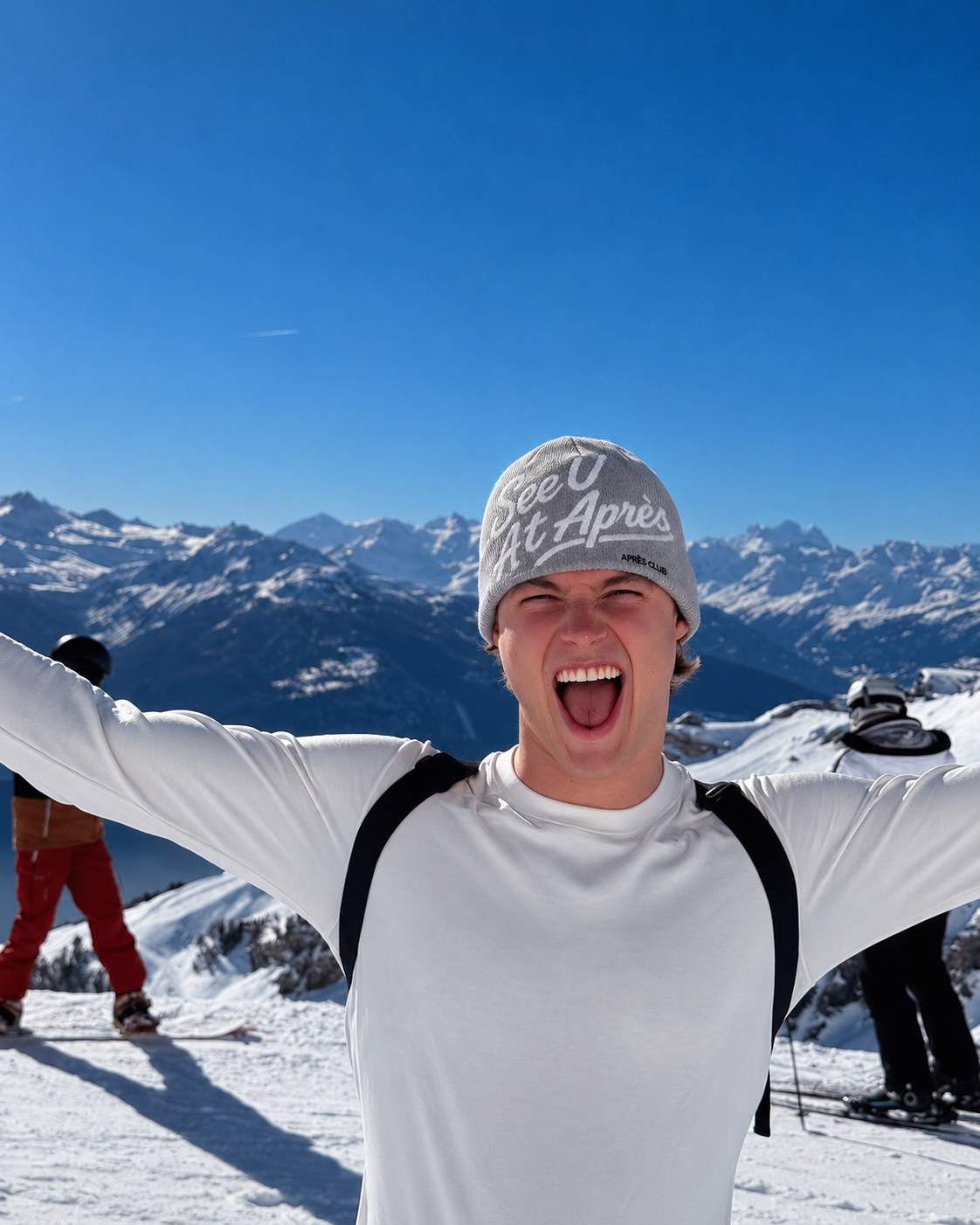 Grey Après Club beanie worn on top of the mountain, arms wide, blue sky behind
