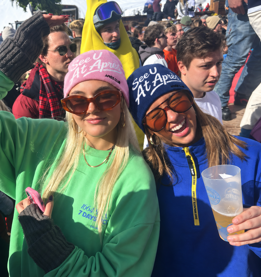 Pink and navy Après Club beanies worn on a packed après terrace